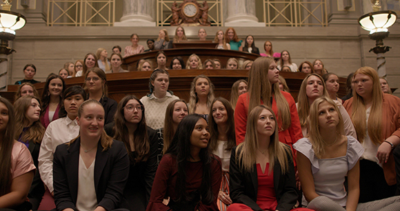 A large group of young women sitting in tiered rows inside a grand hall with architectural details.