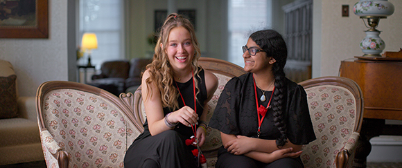 Two young women in black clothing sit on a floral sofa, smiling and looking at each other.