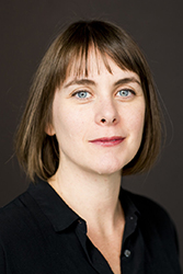 A woman with short brown hair and bangs looks forward against a dark background.