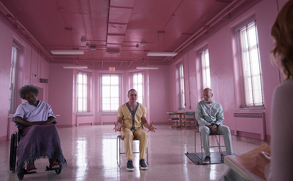 A man in a wheelchair, a man in yellow scrubs, and a man in grey scrubs sit in a large pink room.