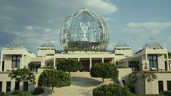 A large, spherical glass dome sits atop a light-colored stone building surrounded by manicured trees and greenery.