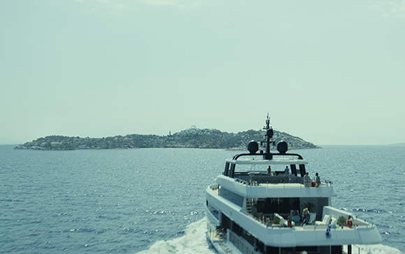 View from behind a large white yacht sailing across blue ocean waters toward a hilly island under a clear sky.
