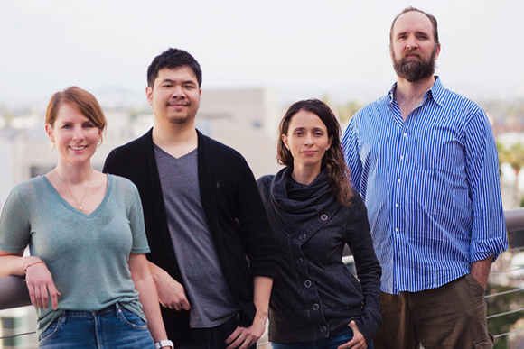 Four people standing in a row on a balcony with a city skyline in the background.