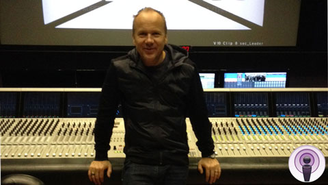 A man in a dark jacket stands behind a large professional audio mixing console in a studio.