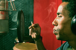 A man in profile exhales smoke next to a studio microphone and pop filter.