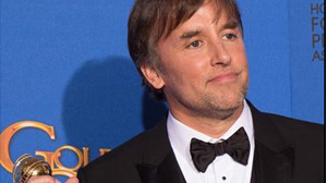 A man in a black tuxedo and bowtie holding a gold award against a blue background.