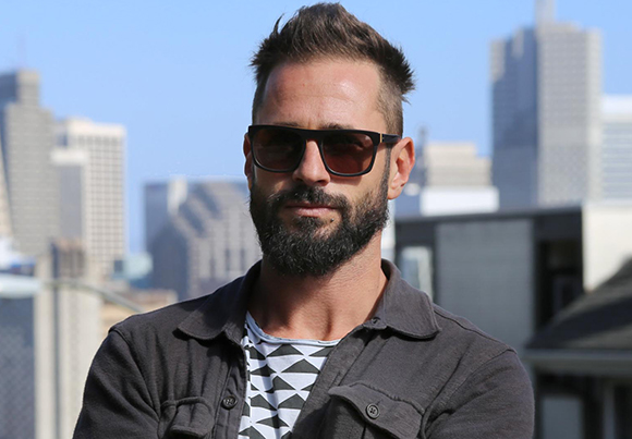 A man with a dark beard and sunglasses stands before a blurred cityscape under a clear blue sky.