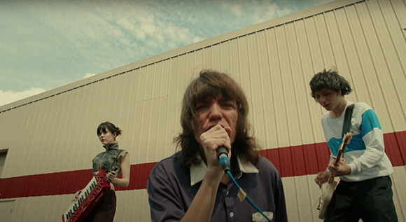 A singer with a microphone, a keyboardist, and a guitarist performing outdoors in front of a tan warehouse.