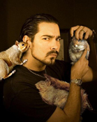 A man with dark hair and a goatee holds a small rodent while two squirrels rest on his shoulder.