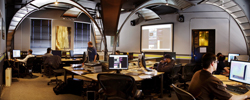 People working at computer desks in a large office with curved metal ceiling structures.