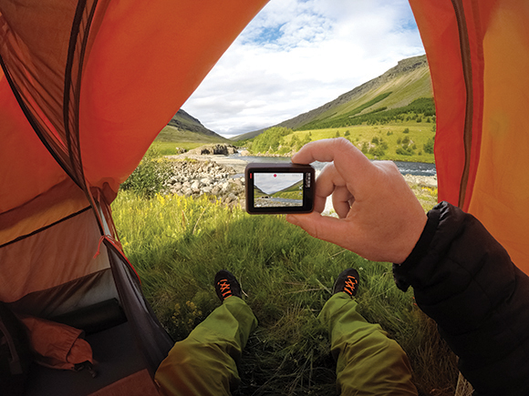 A person holds a digital camera inside an orange tent, framing a view of a green valley and river.