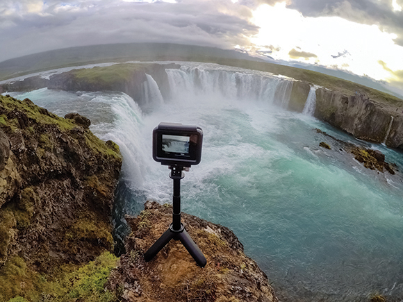 A digital camera on a small tripod sits on a rocky ledge overlooking a wide, powerful waterfall.