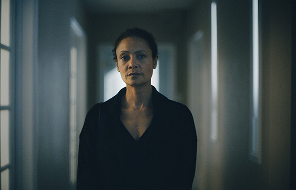 A woman with dark hair pulled back stands in a dark, narrow hallway looking directly at the camera.