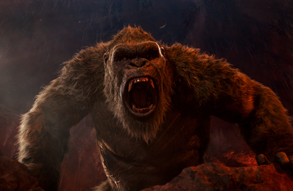 A large, muscular gorilla with dark fur roars with its mouth wide open against a dark, smoky background.