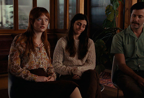 A woman with red hair looks up while two others sit beside her in a dimly lit room.