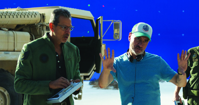 A man in a green jacket talks to a man in a blue shirt and white cap in front of a blue screen.