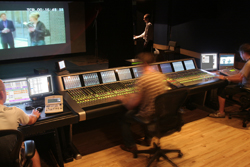Technicians operating large audio mixing consoles in a dark control room with monitors.