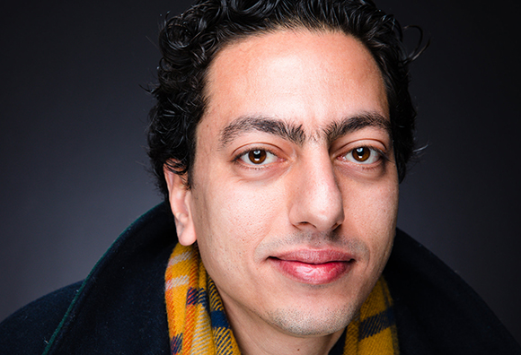 Close-up portrait of a man with dark curly hair and brown eyes wearing a yellow patterned scarf.