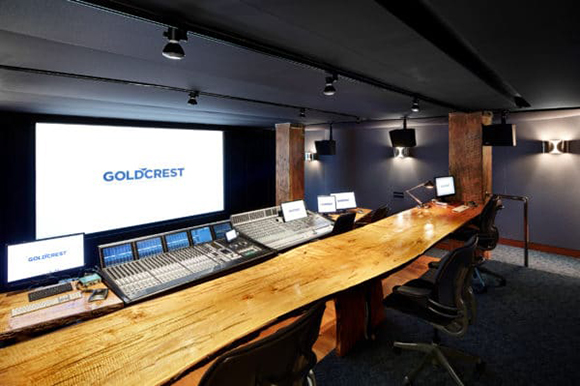 A dark recording studio featuring a long live-edge wooden desk, mixing consoles, and a large screen.