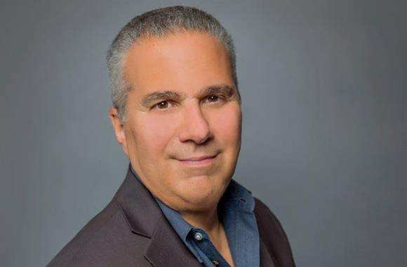 A middle-aged man with short graying hair wearing a dark blazer and blue shirt against a gray background.