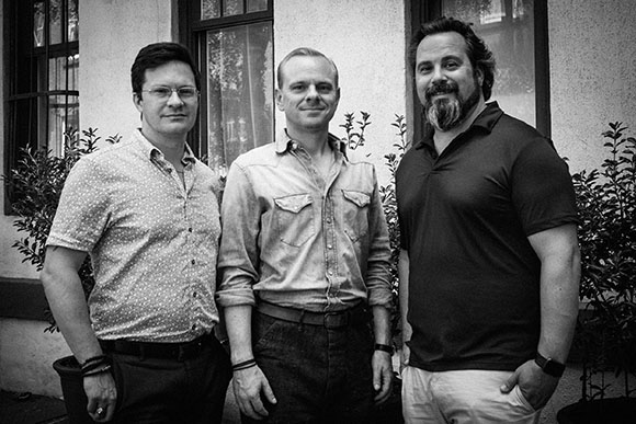 Three men standing side by side in front of a building with windows and plants.
