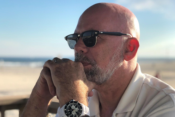 A bald man with a grey beard and sunglasses rests his chin on his hands while looking toward the ocean.