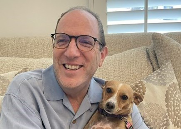 A smiling man wearing glasses sits on a couch and holds a small brown and white dog.