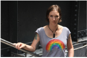 A woman with short brown hair wearing a grey t-shirt with a rainbow graphic, standing by a metal railing.