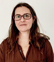 A woman with long brown hair and glasses wearing a brown button-down shirt against a white background.