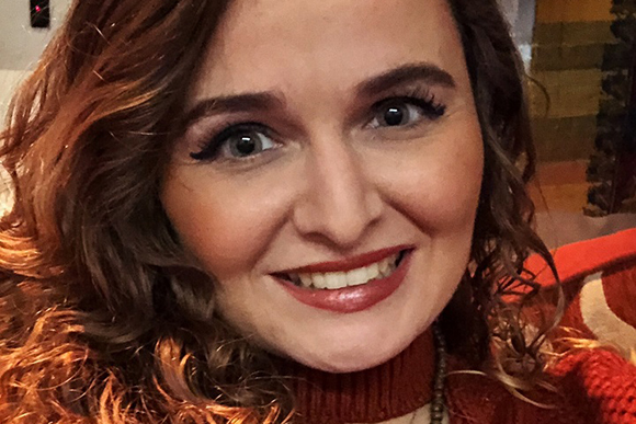 A smiling woman with wavy reddish-brown hair and dark eye makeup looking toward the camera.