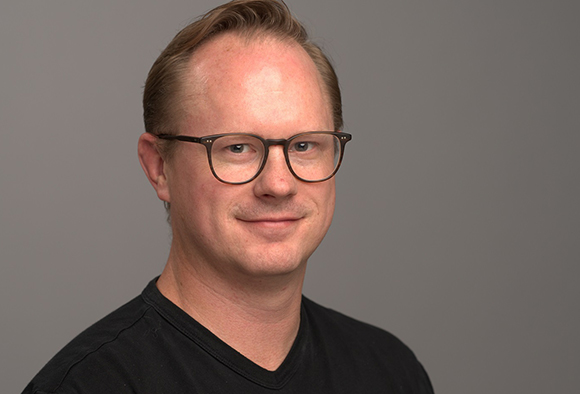 A man with light hair and black-rimmed glasses smiling against a gray background.