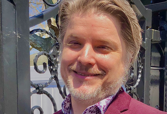 A smiling man with light brown hair and a beard wearing a maroon jacket and patterned shirt.