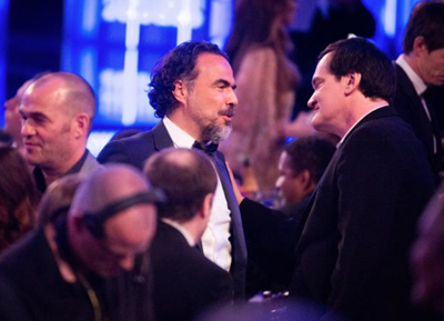 Two men in suits talk to each other at a crowded indoor event with blue lighting.