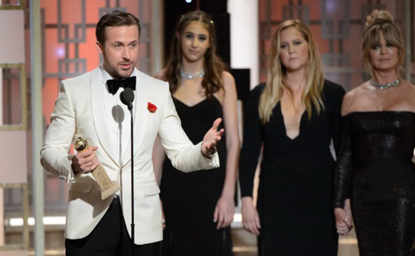 A man in a white tuxedo holds a gold award and speaks into a microphone on stage.