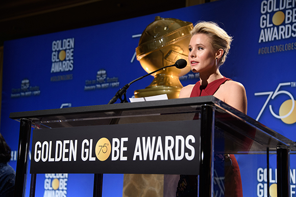 A woman in a red dress speaks at a podium with Golden Globe Awards branding in front of a large gold trophy.