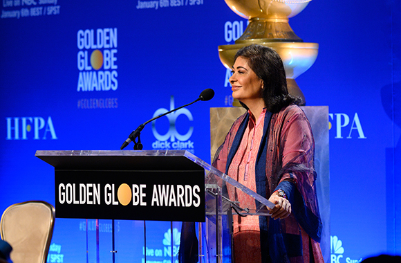 A woman in a pink patterned shirt and blue shawl stands behind a Golden Globe Awards podium.