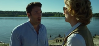 A man in a light blue shirt looks at a woman with blonde hair by a large body of water.