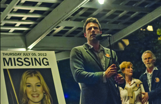 A man in a grey blazer speaks into a microphone beside a large missing person poster of a blonde woman.