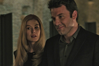 A man in a dark suit and a blonde woman in black stand close together in a dimly lit room.
