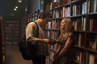 A man with a backpack and a woman in a patterned dress talk in a dimly lit library aisle.