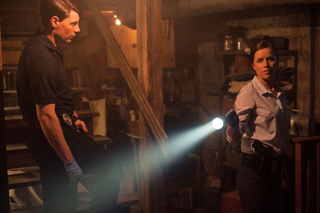 A man and a woman stand in a dark, cluttered basement, each holding a flashlight.