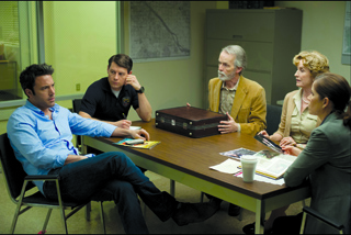 Five people sit around a wooden conference table in an office, discussing a dark briefcase.