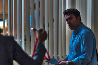 A man in a blue button-down shirt looks toward a person holding a red object in front of vertical blinds.