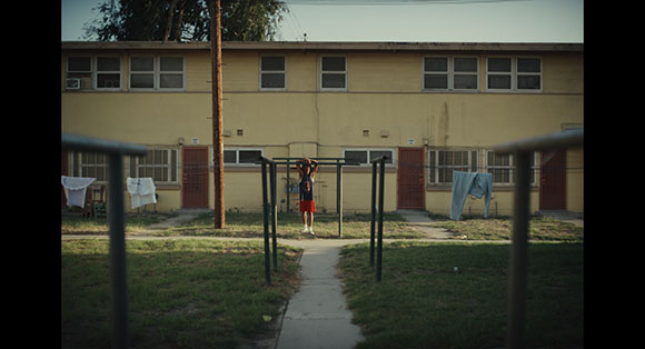 A person hangs from a pull-up bar on a grassy area in front of a long, two-story apartment building.