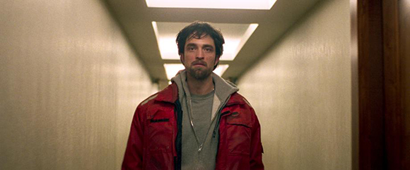 A man with dark hair and a beard wearing a red jacket and grey hoodie stands in a brightly lit hallway.