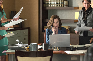 A woman in a blue blazer sits at a desk with a laptop while others hold documents in an office.