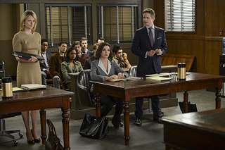 Professionals in a courtroom with a woman in a tan dress standing and a man in a suit standing near a desk.