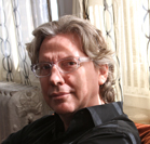 A man with wavy light brown hair and glasses wearing a black button-down shirt.