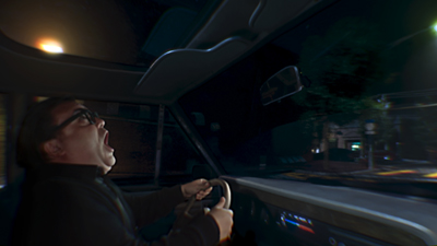 A man with glasses driving a car at night, looking up with a wide-mouthed, shocked expression.