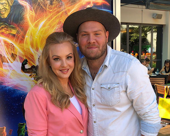 A blonde woman in a pink blazer and a man in a brown hat and light blue shirt smiling together.